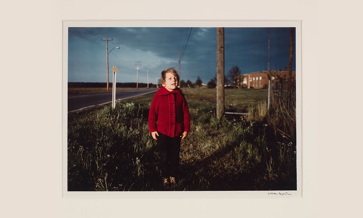 Photo of a little boy wearing a red sweater standing in the grass but the side of a road