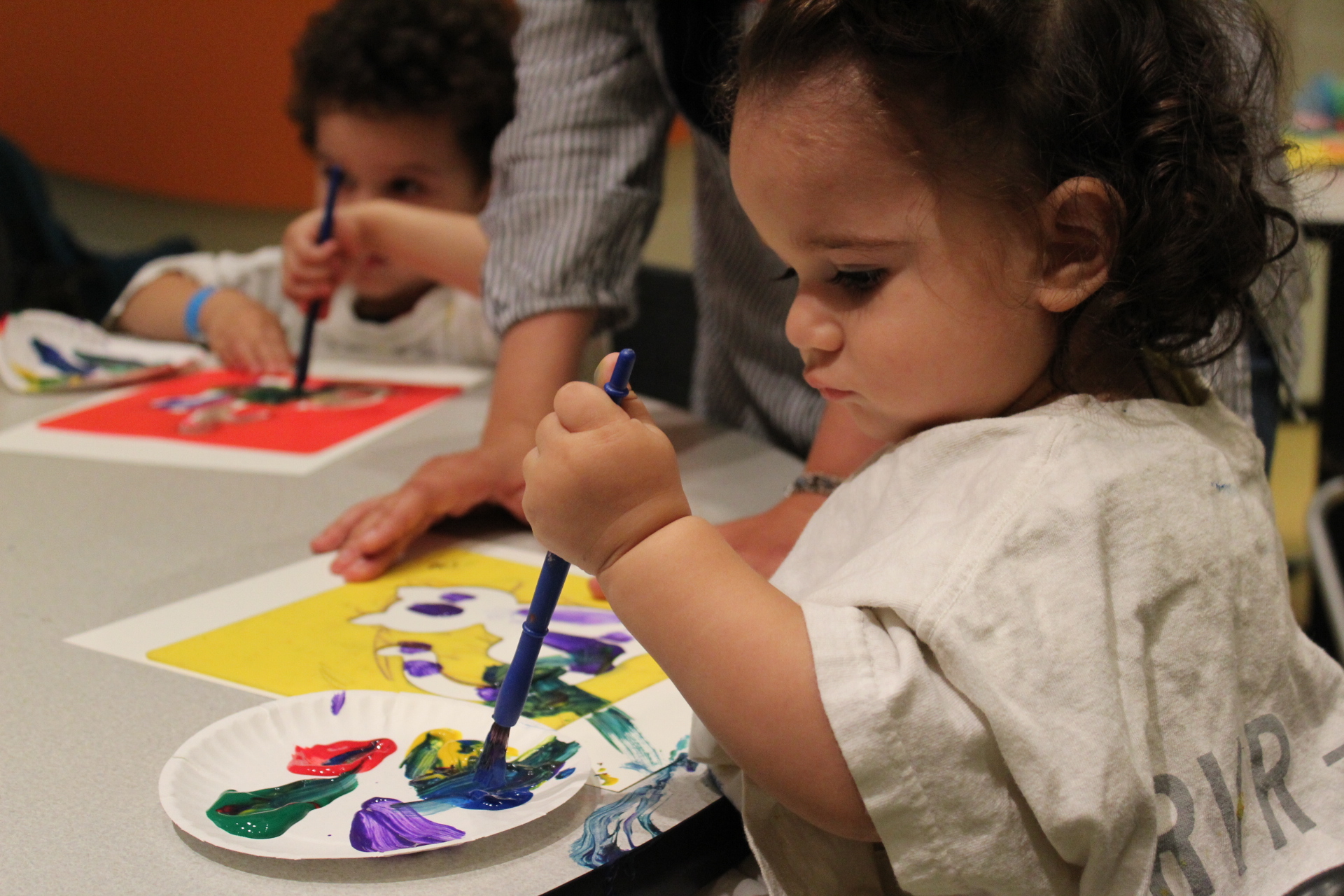Young child wearing a smock, dips paintbrush into paint on a small paper plate