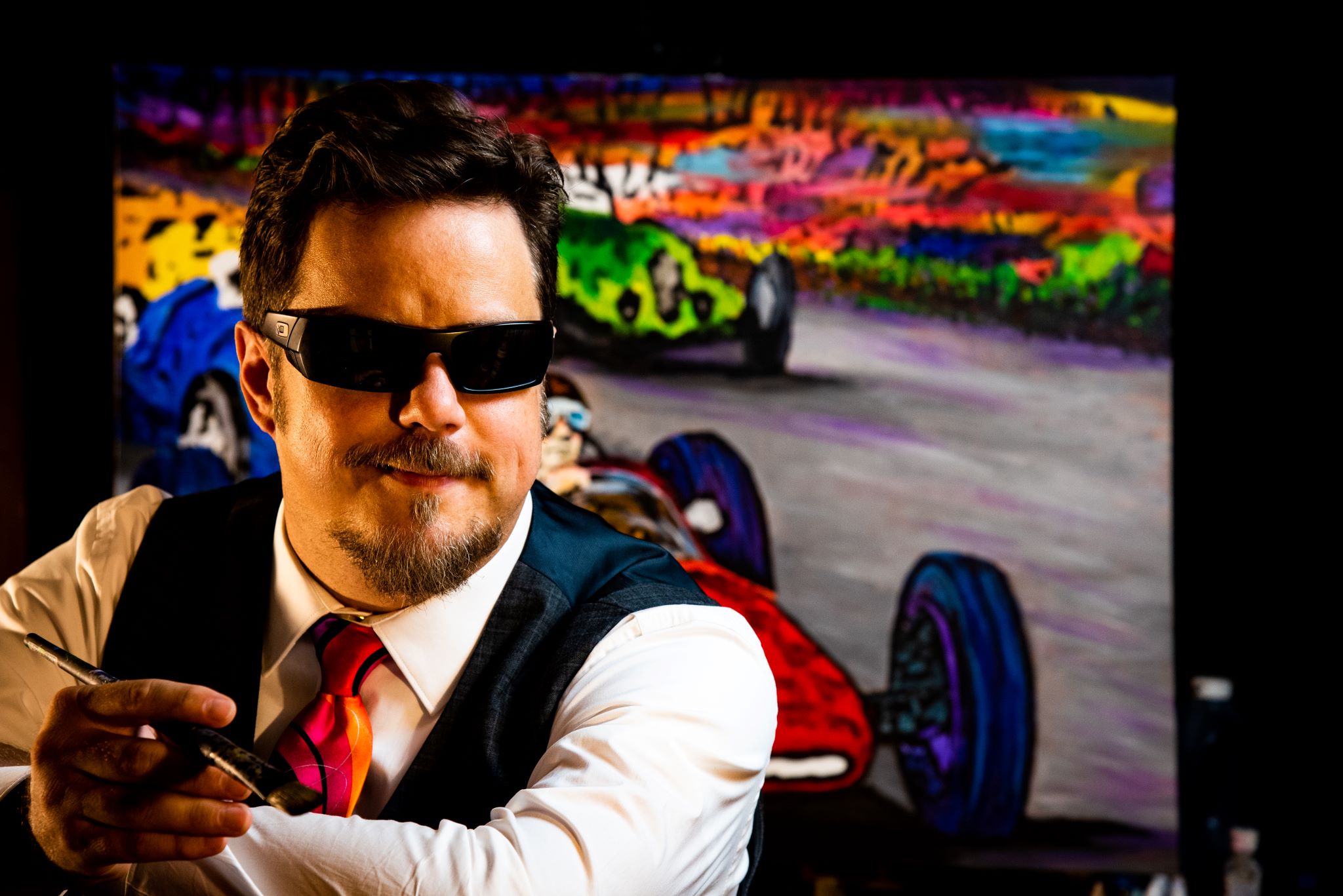 John Bramblitt sits, holding a paintbrush, in front of a colorful painting of race cars