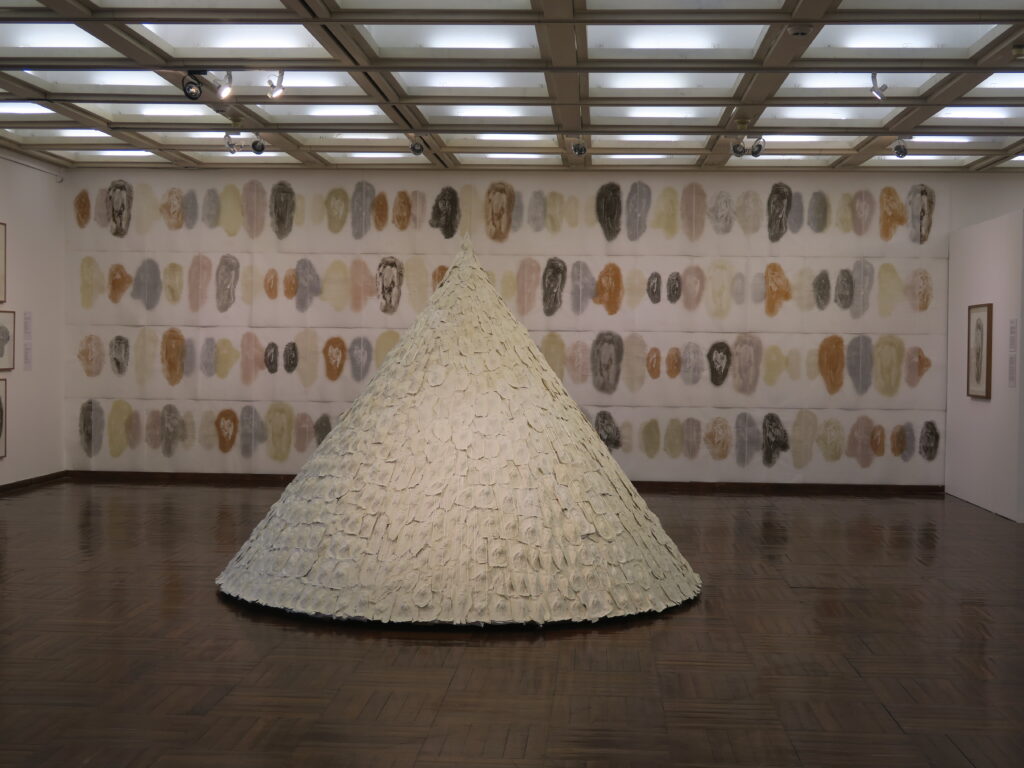 White conical shaped installation sitting on floor with prints behind it on a wall