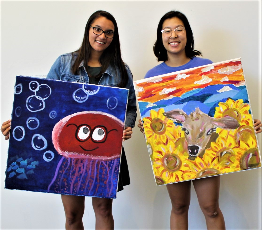 Two people hold their own ceilings tiles. One ceiling tile depicts a jellyfish and bubbles on a dark blue background. The other ceiling tiles depicts a baby cow's face peeking our from a bunch of sunflowers with a sunset in the background.