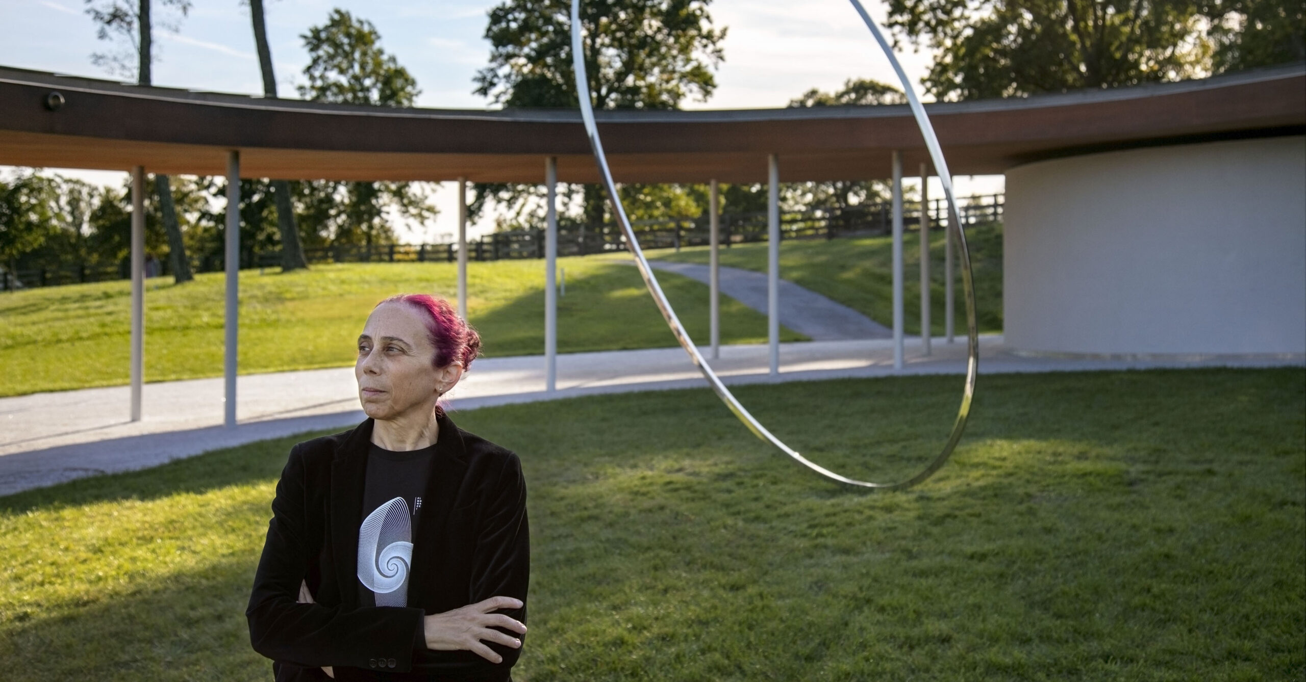 A woman standing in a sculpture garden with a large silver circular sculpture behind her