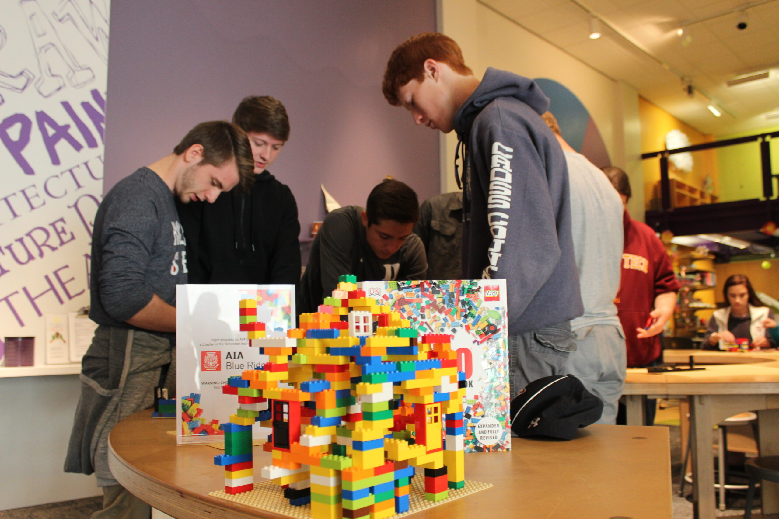 A building created from Legos stands on a table in the foreground of the picture as 4 teenagers actively build their owns Lego sculptures