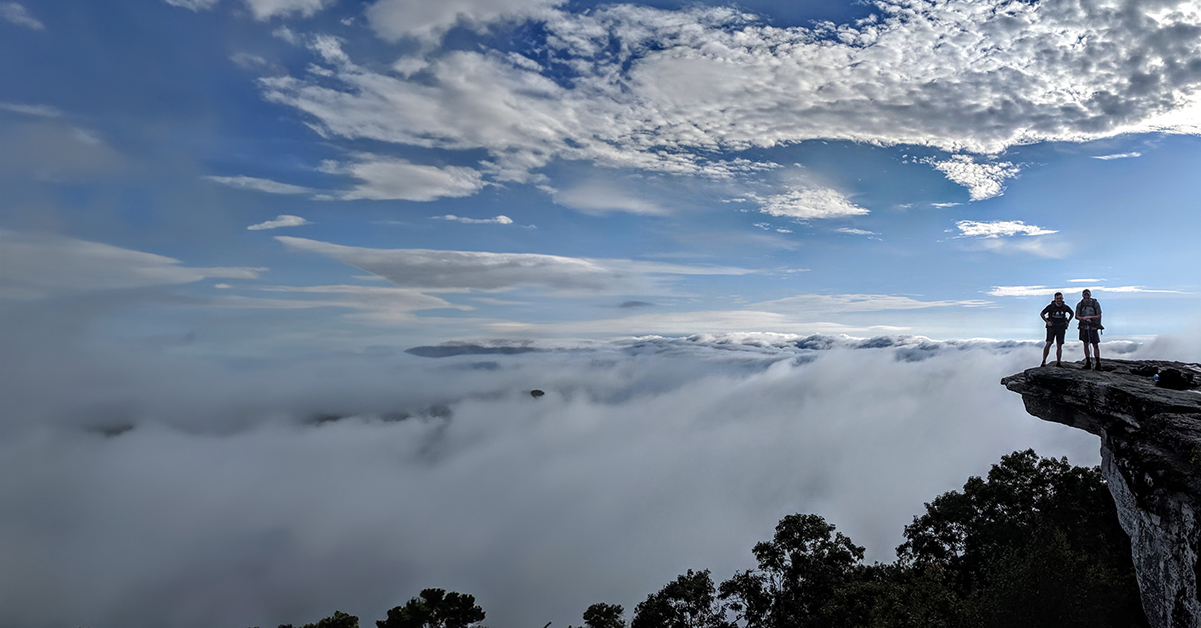 Two people standing on a mountain outcrop with clouds above and trees below