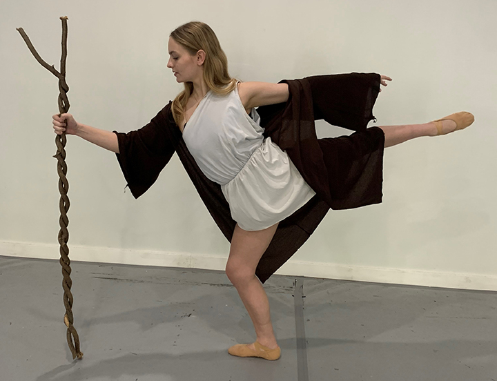 A woman in a white dress and black jacket holds on to a tall stick while performing a ballet move