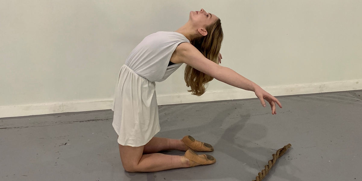 A woman in a white dress kneels on the floor and arches her arms and head backward while she looks up