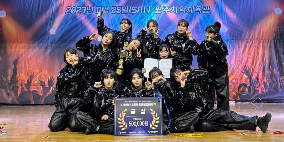 A K-Pop group standing together with a banner in Korean at their feet