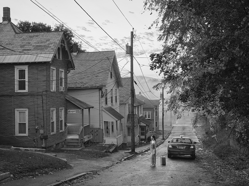 Black and white photo of a woman standing near a taxi and a row of clapboard houses