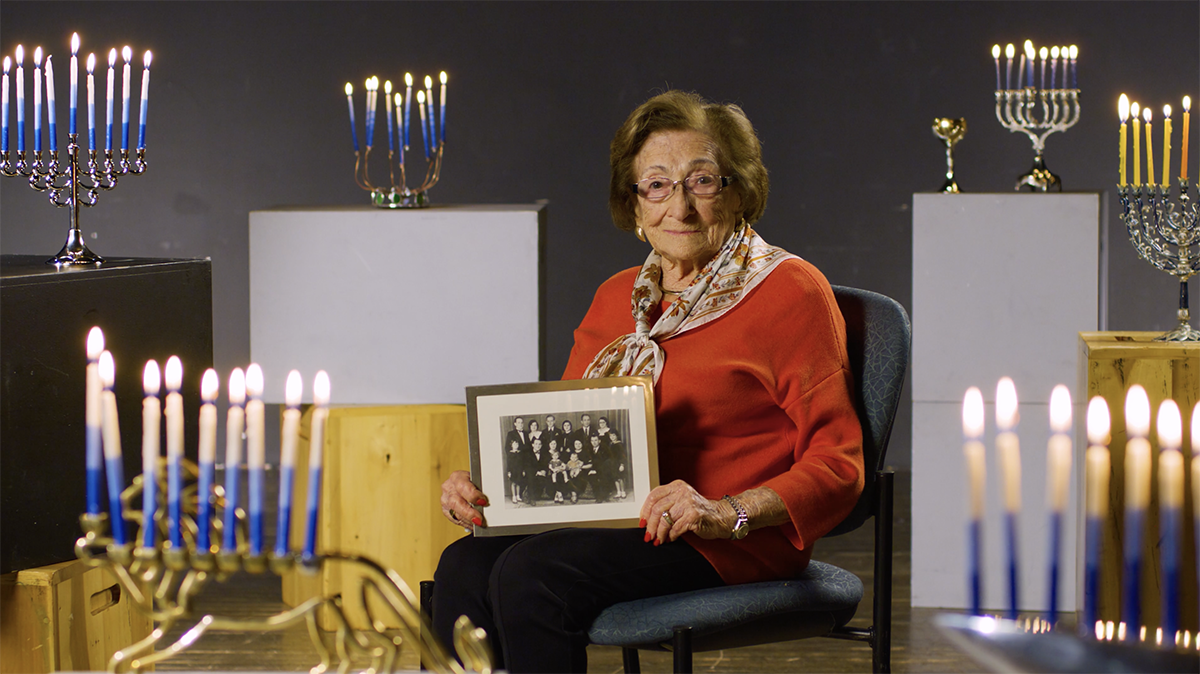A woman sits in a chair holding a framed photo while surrounded by lit menorahs