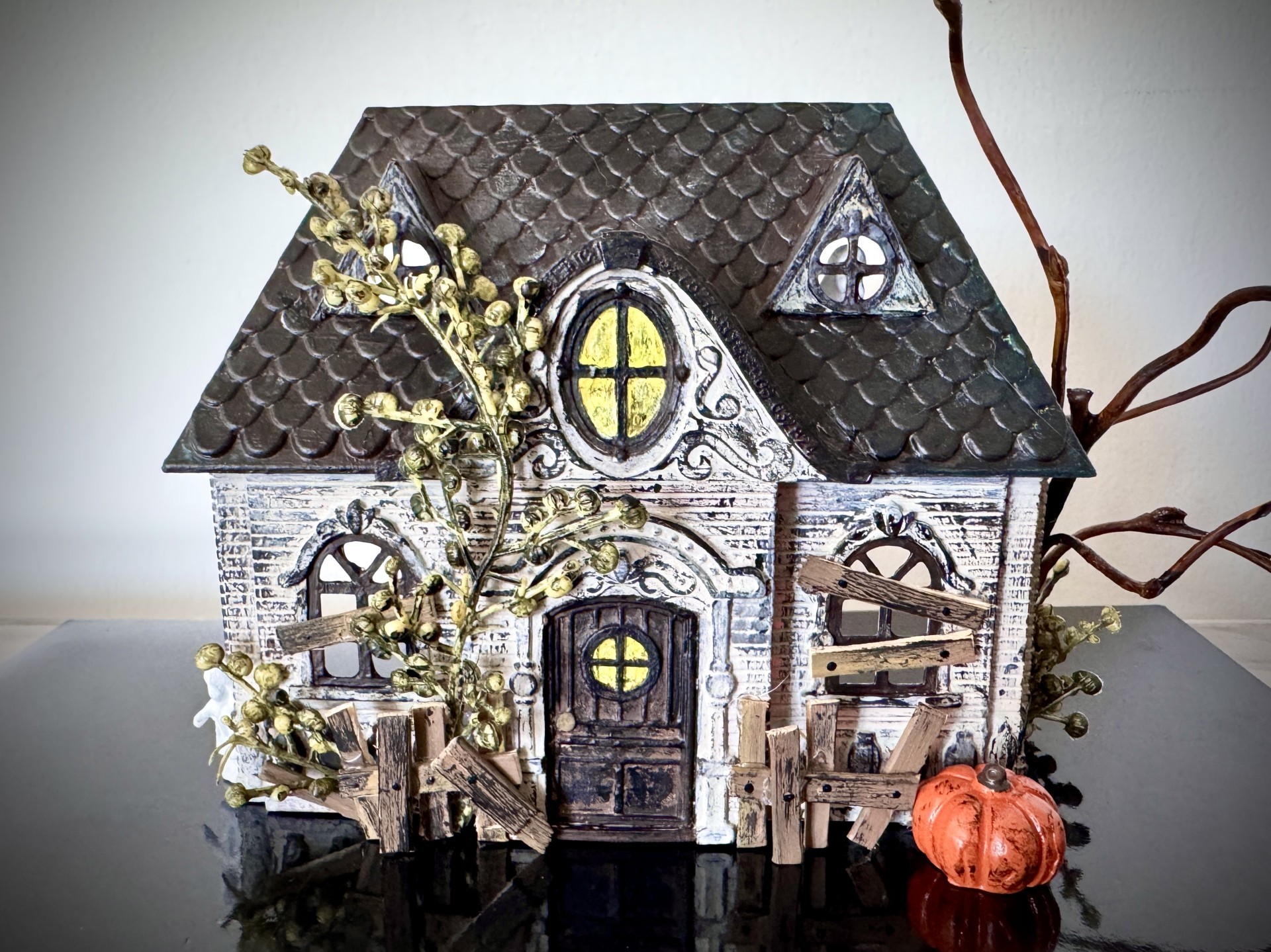 Miniature haunted house is displayed with broken fence, feaux greenery, and tiny pumpkin