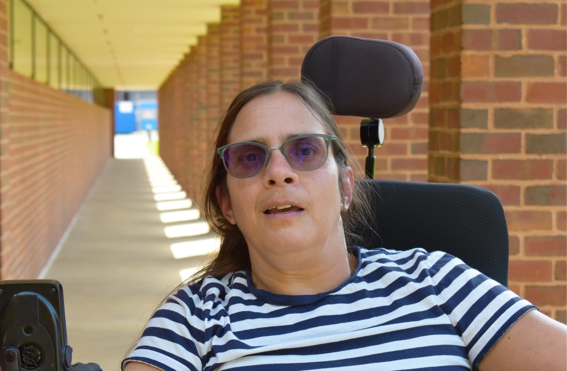 A woman in sunglasses smiles while she sits in a wheelchair with a long corridor of brick columns extending behind her