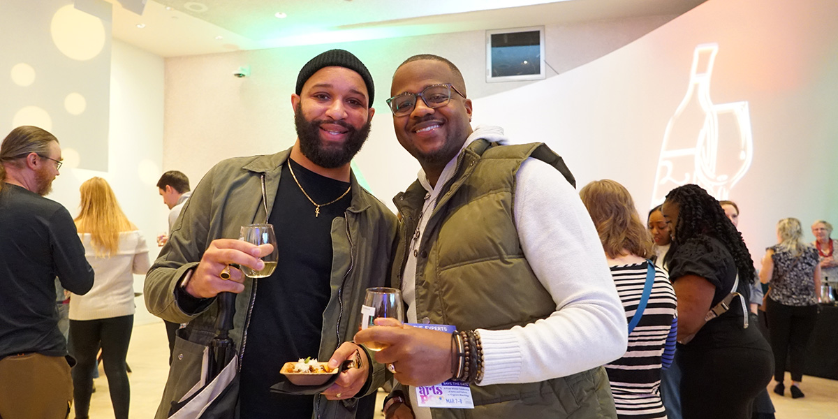 Two men hold glasses of wine while standing in a crowd