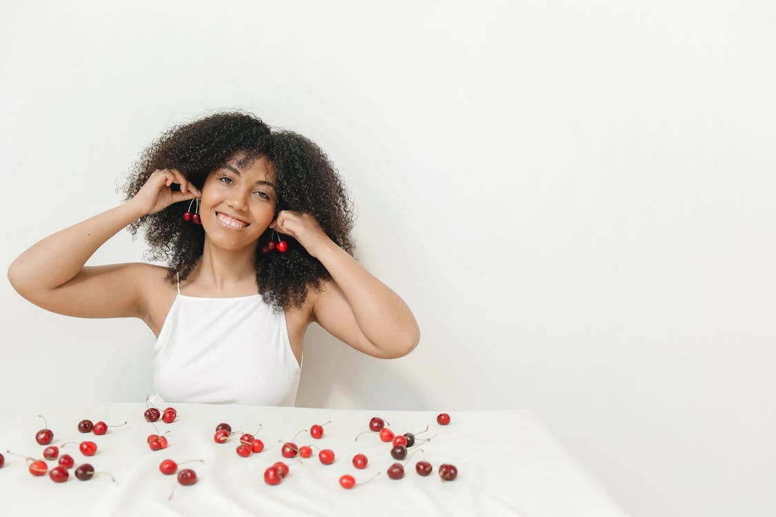 A woman sits behind a table with cherries spread on it and holds cherries up to her ears like earrings