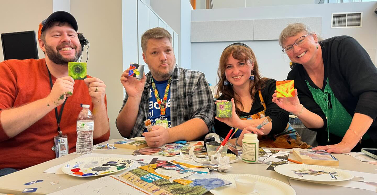 Four adults sit at a table covered in art-making supplies and hold up small canvases that they painted.