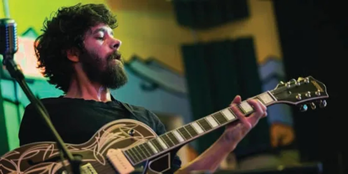 A man with a brown beard and black t-shirt plays a guitar with a brightly colored mural wall behind him
