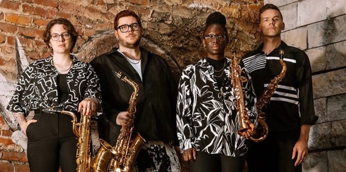 Four people stand against a brick wall holding brass instruments