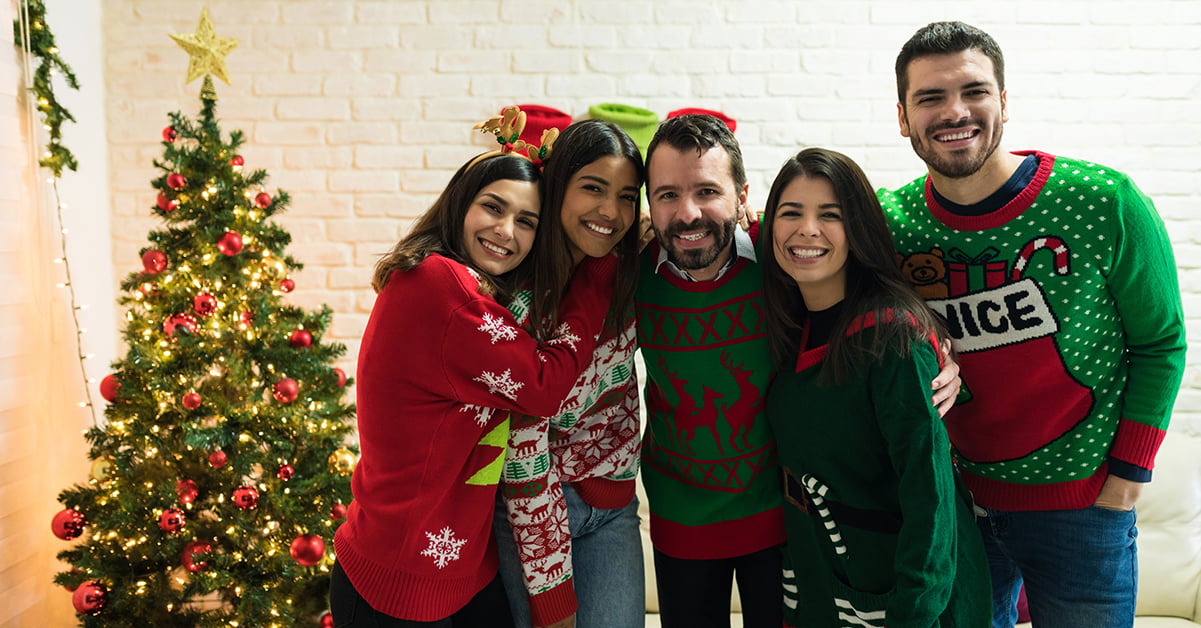 Four people dressed in green and red holiday sweaters smile near a Christmas tree