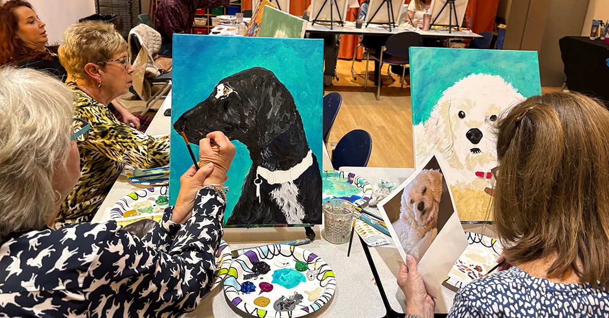 A group of women sit at adjoining tables and paint canvases featuring portraits of dogs